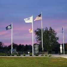 Flags of the United States and other banners fly at dusk. Green grass and a memorial are in view.