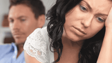 Woman with sad expression, hand on forehead. Man in background, both appear unhappy.