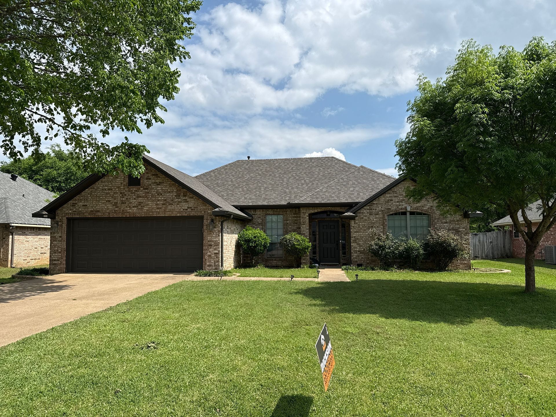 Suburban brick house with a two-car garage, front lawn, and trees under a partly cloudy sky