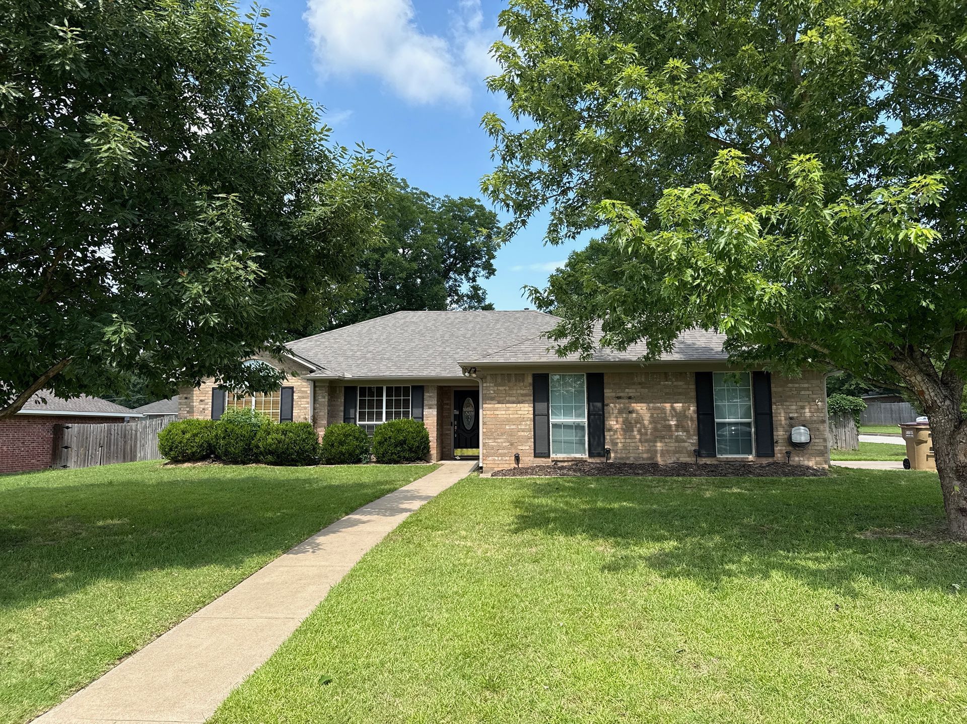 Single-story brick house with a front lawn, sidewalk, and large trees under a blue sky