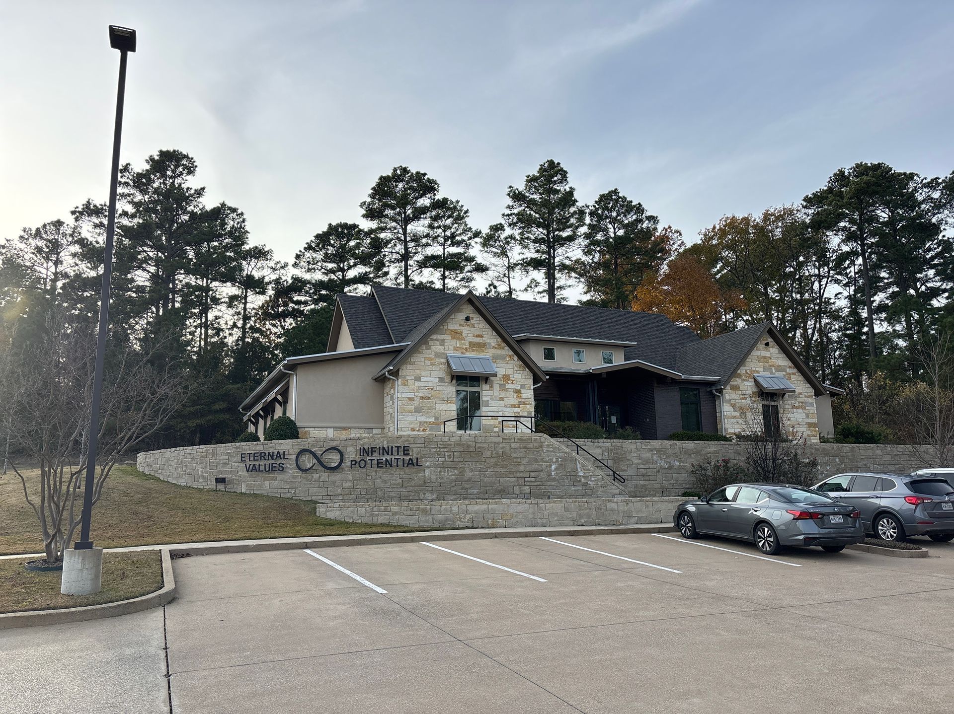 Stone building with dark roof behind a retaining wall, with parked cars in a lot and trees in the background.
