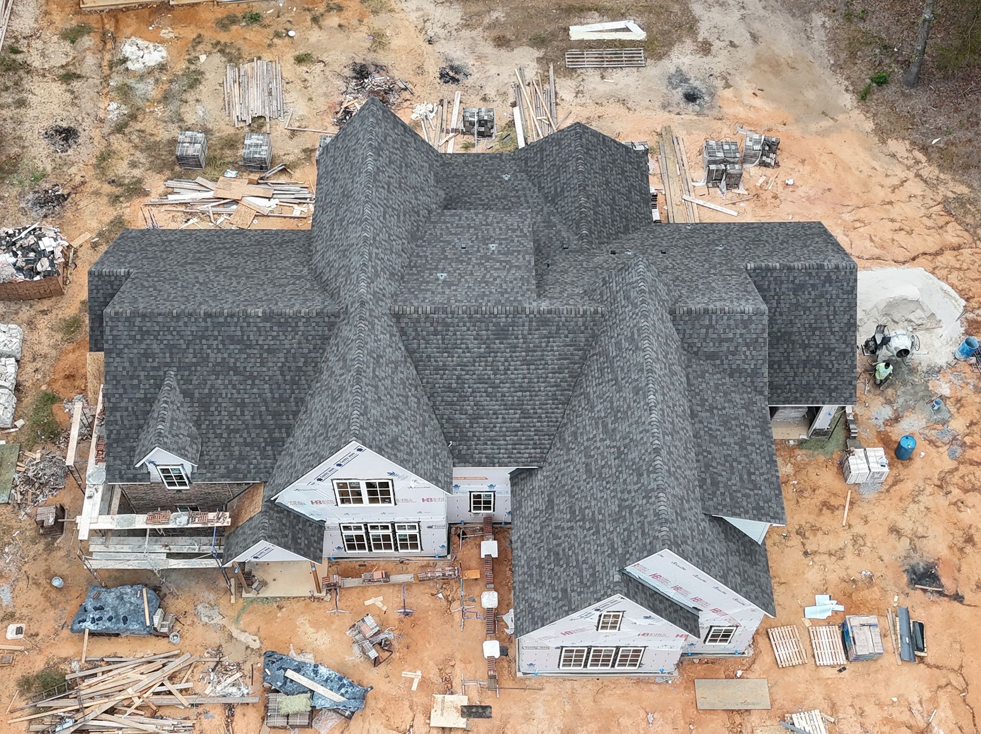 Aerial view of a large house with dark roof under construction, surrounded by building materials and dirt.