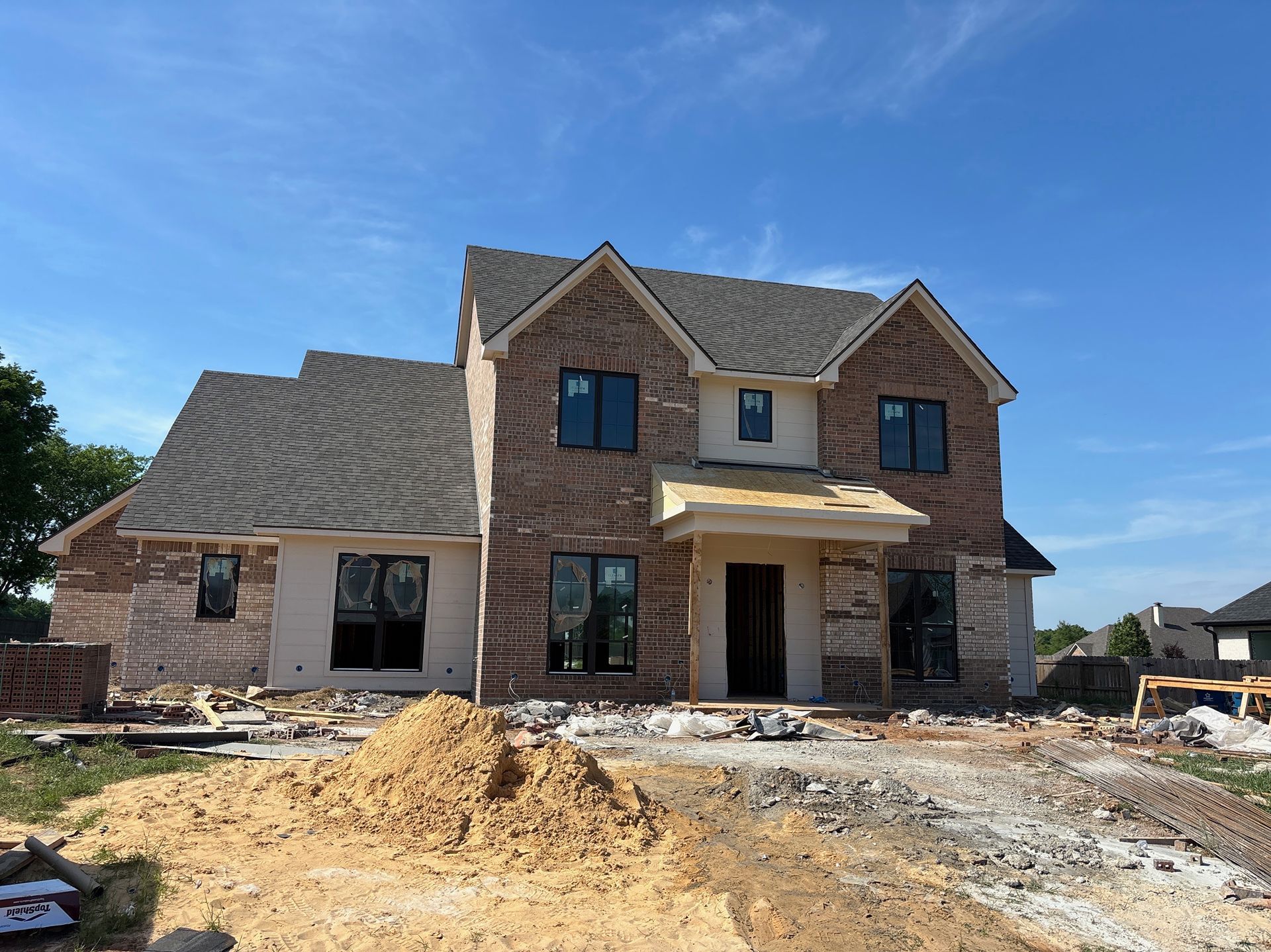 Partially built brick house with roof shingles and dirt driveway under a blue sky