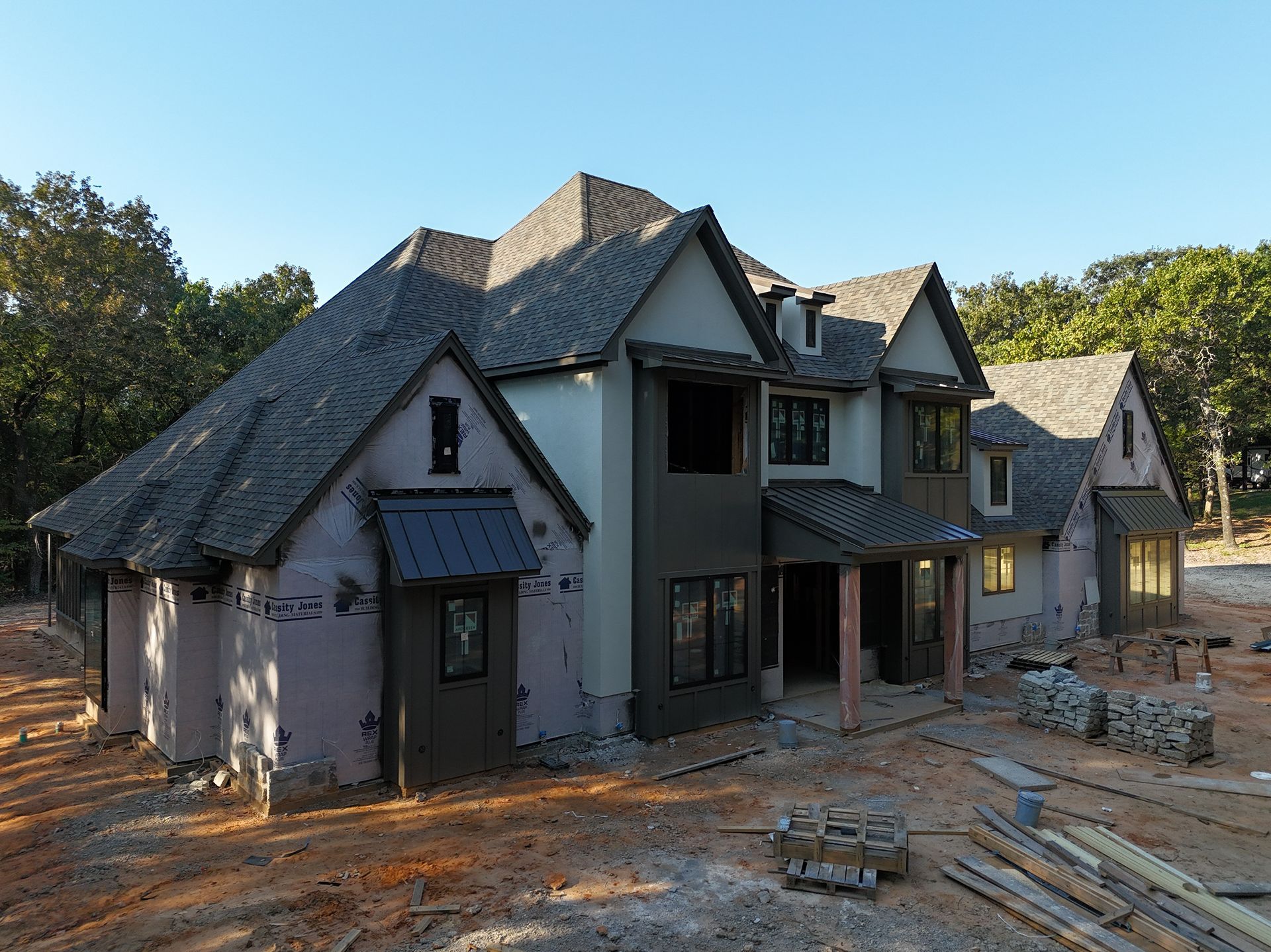 Large unfinished house with gray roof and stucco walls at a construction site, surrounded by materials and dirt.