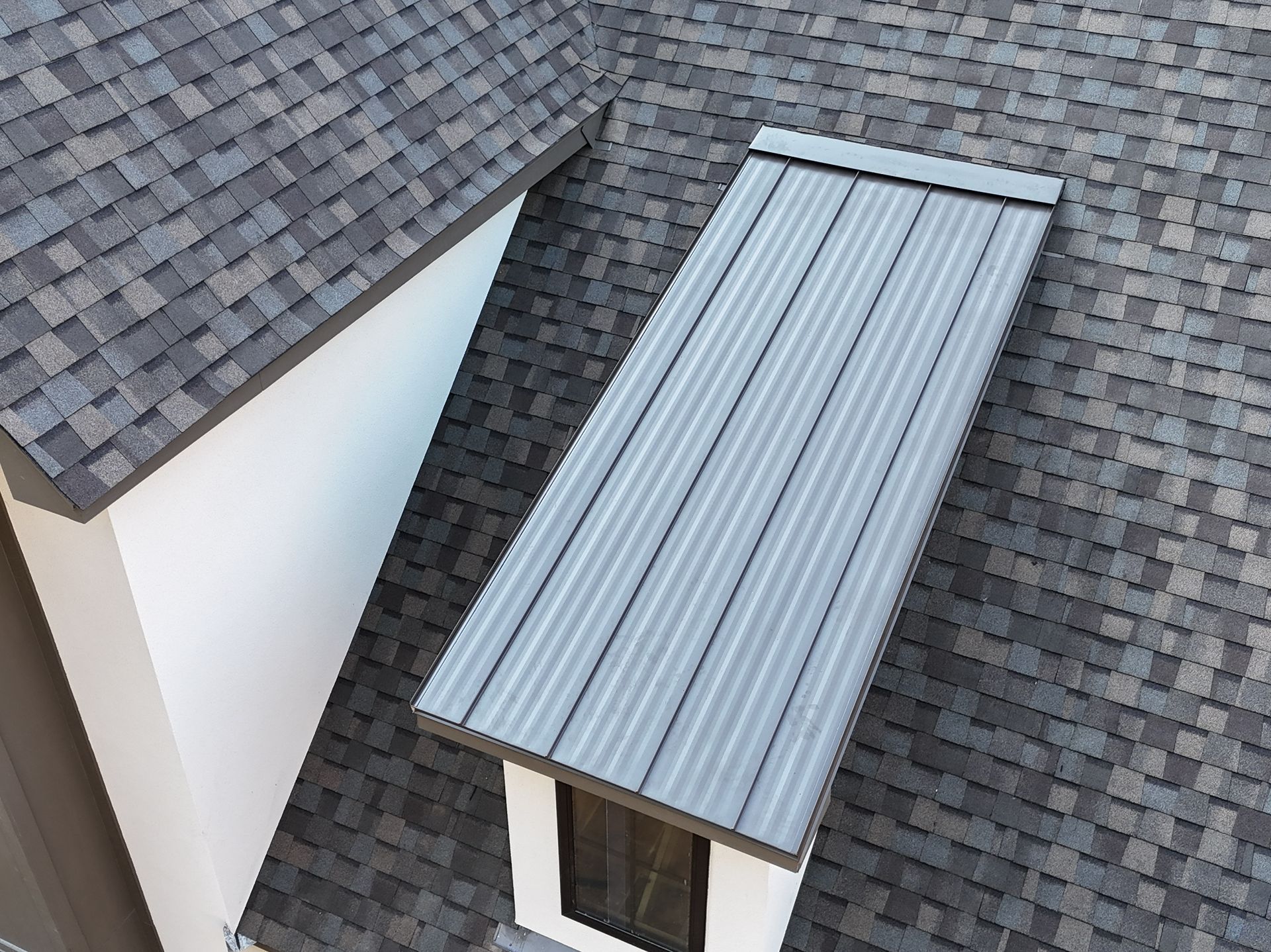 Roofing with gray shingles and a light metal awning between two house roofs.