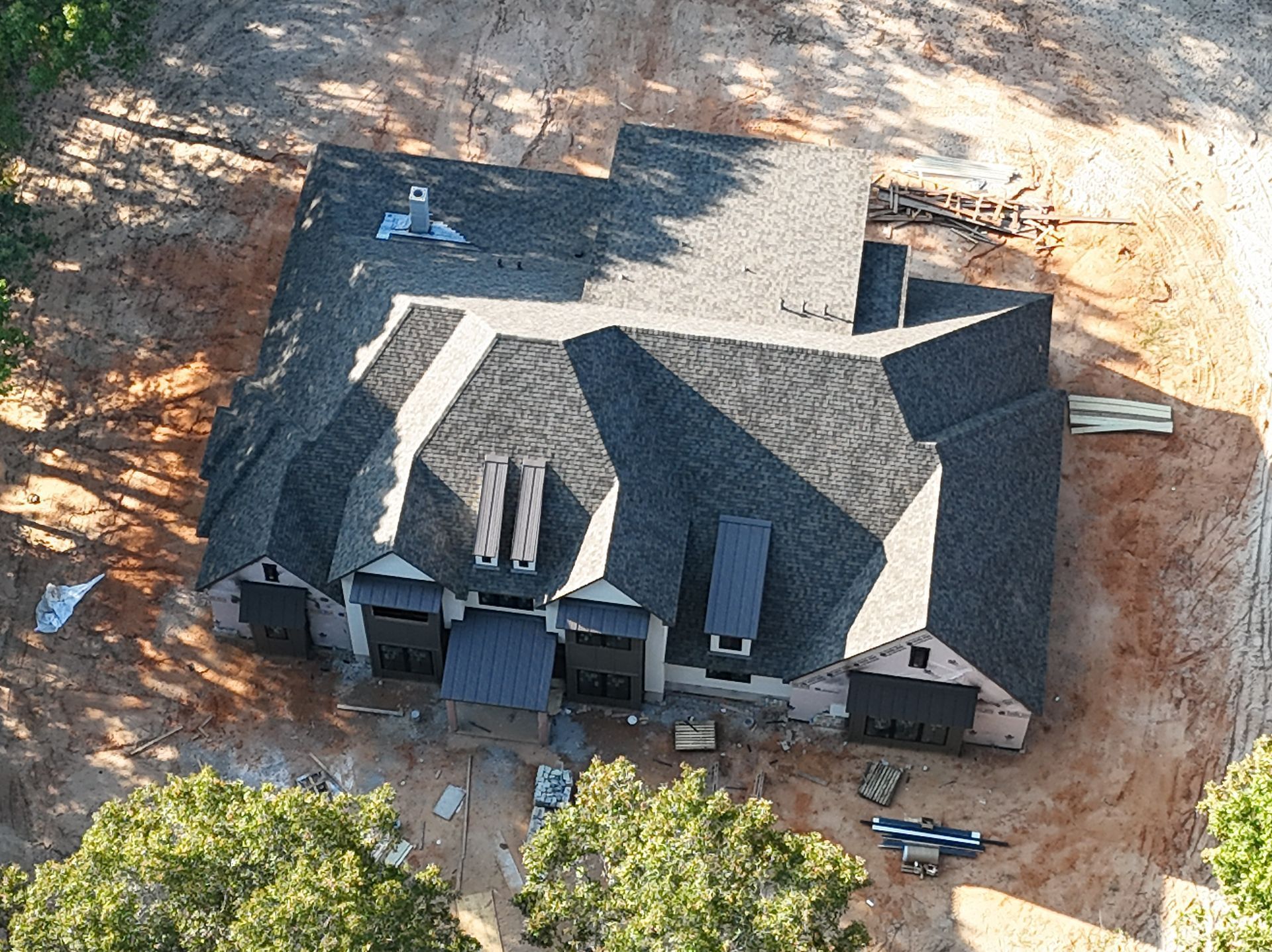 Aerial view of a large house with a dark roof under construction on a dirt lot.