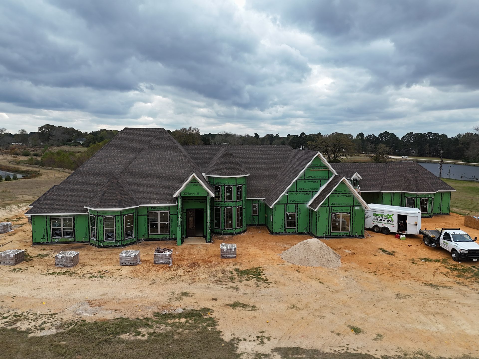 Green house under construction on a dirt lot beneath a cloudy sky, with a white truck nearby.