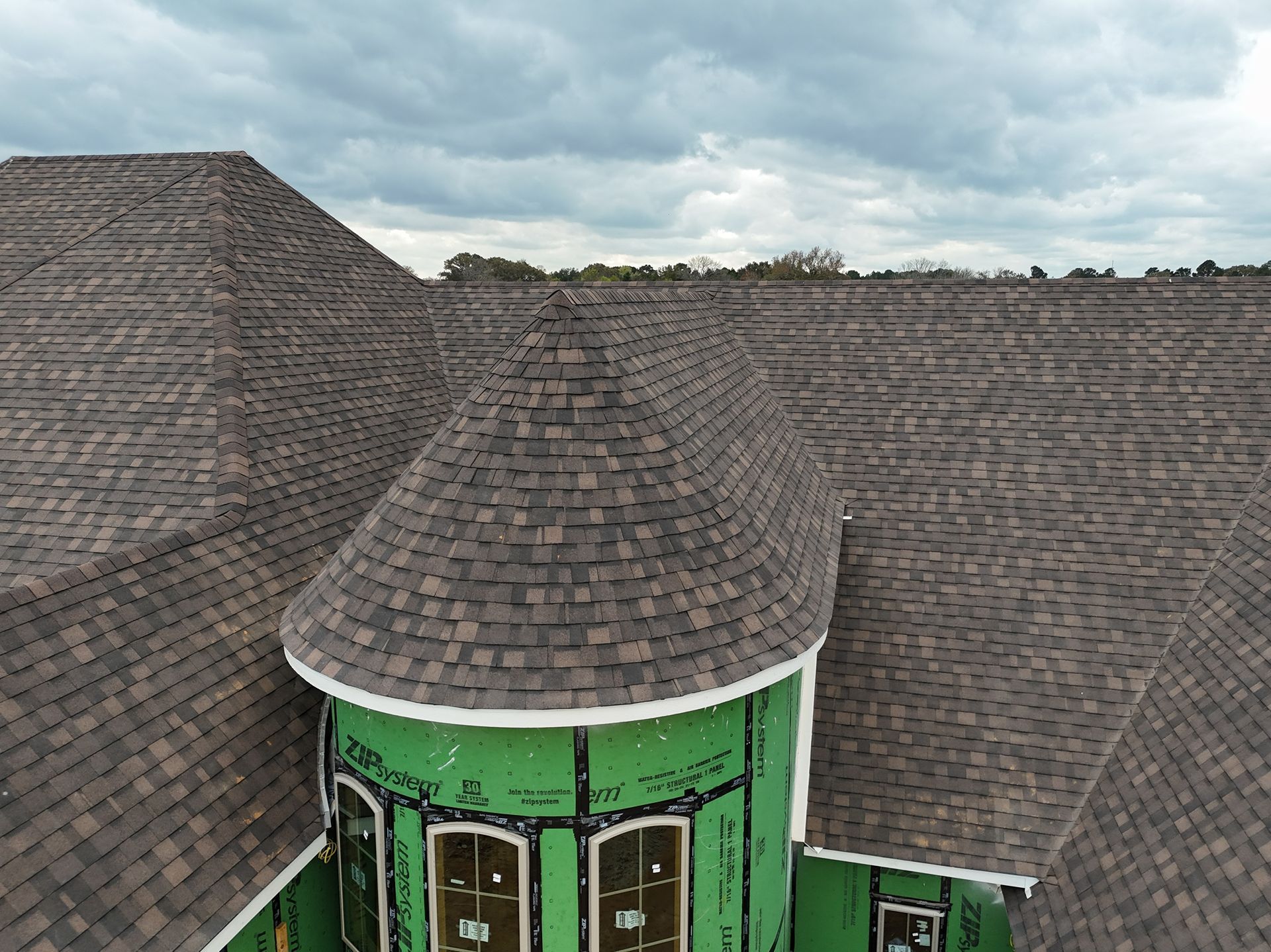 Aerial view of a green turret roof with dark shingles on a multi-roof building under a cloudy sky