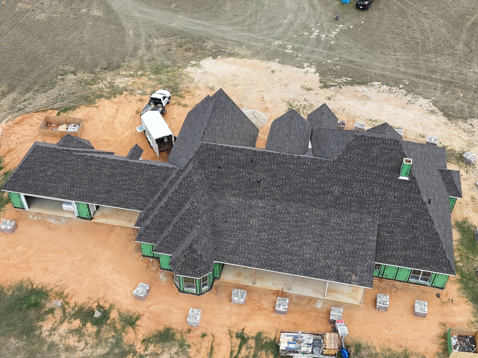 Aerial view of a large dark-roofed house under construction on a dirt lot, with workers and vehicles nearby