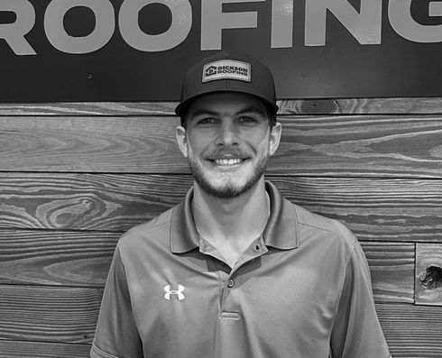Smiling man in a cap and polo standing before a wood wall with a roofing sign.