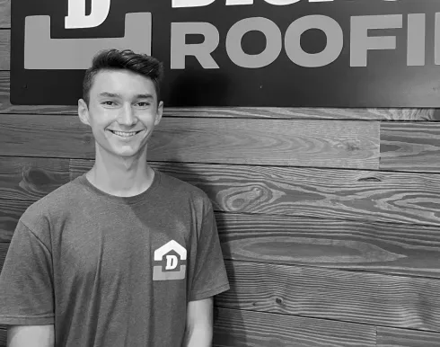 Smiling man in a gray shirt stands indoors before a wood wall and a roofing sign.