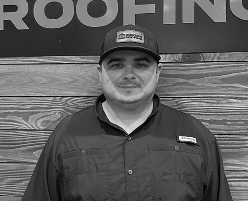 Man standing in front of a wood wall with a partially visible roofing sign, wearing a cap and dark shirt