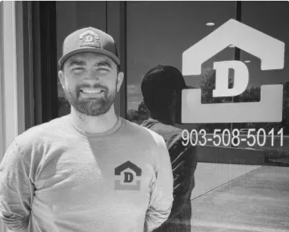 Smiling man in cap and T-shirt stands beside a glass door with a logo and phone number 903-508-5011