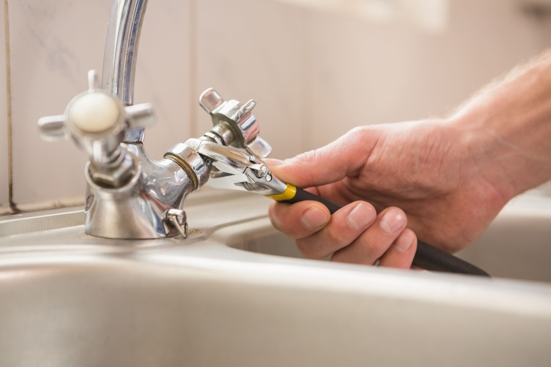 Hand using a wrench to repair a silver faucet in a stainless steel sink.