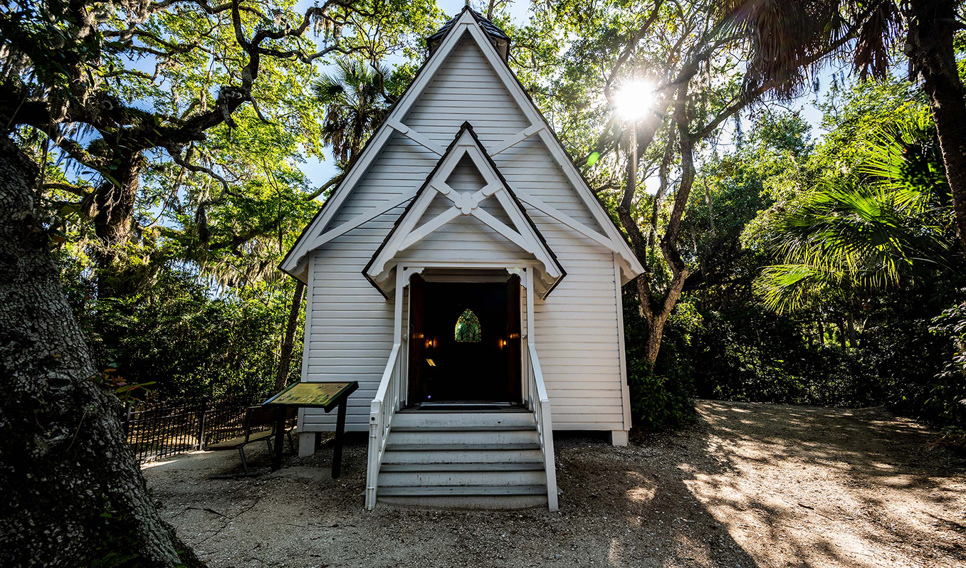 A small white church in the middle of a forest with the sun shining through the trees.