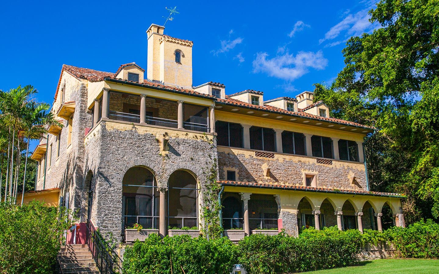 A large stone building with arches and stairs is surrounded by trees and bushes.