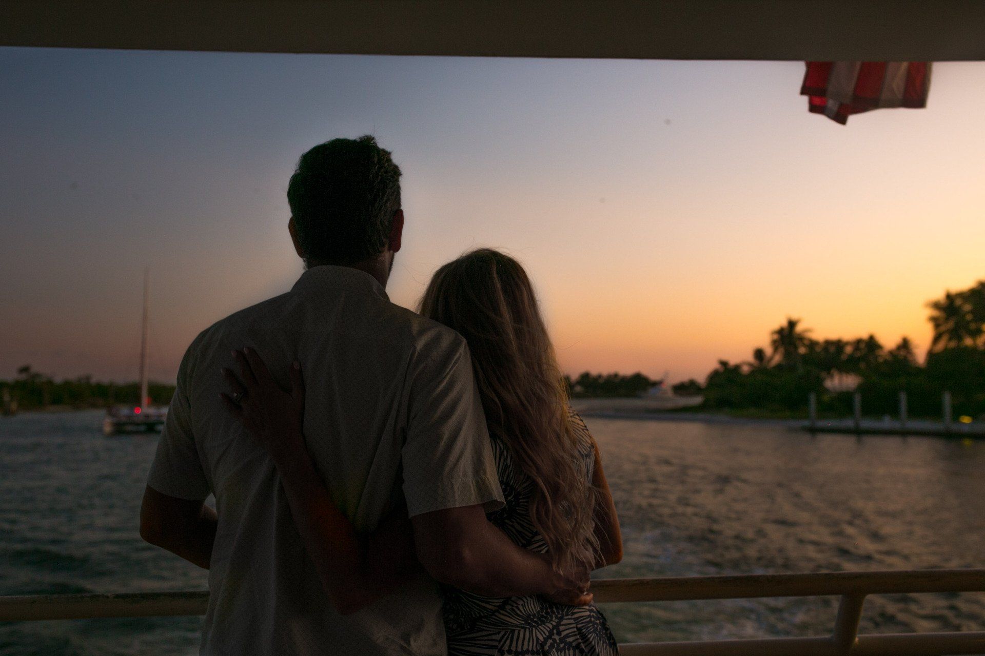 A man and a woman are standing on a boat looking at the water at sunset.