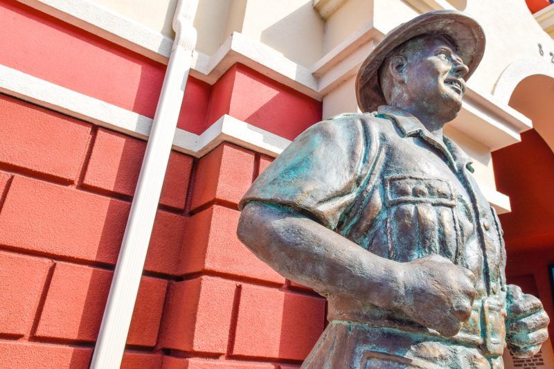 A statue of a man in a hat is standing in front of a red brick building.