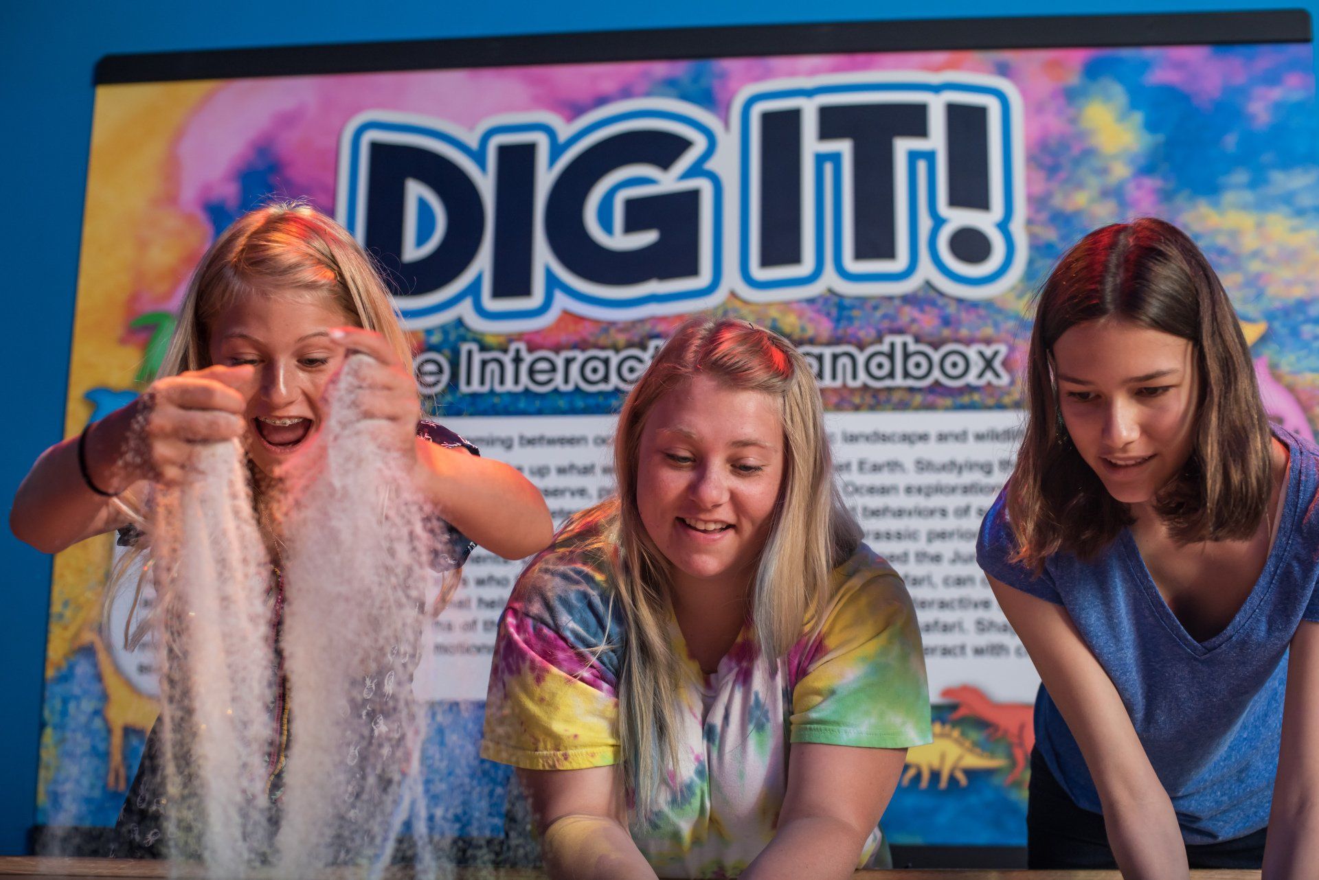 Three girls are playing with sand in front of a sign that says dig it !