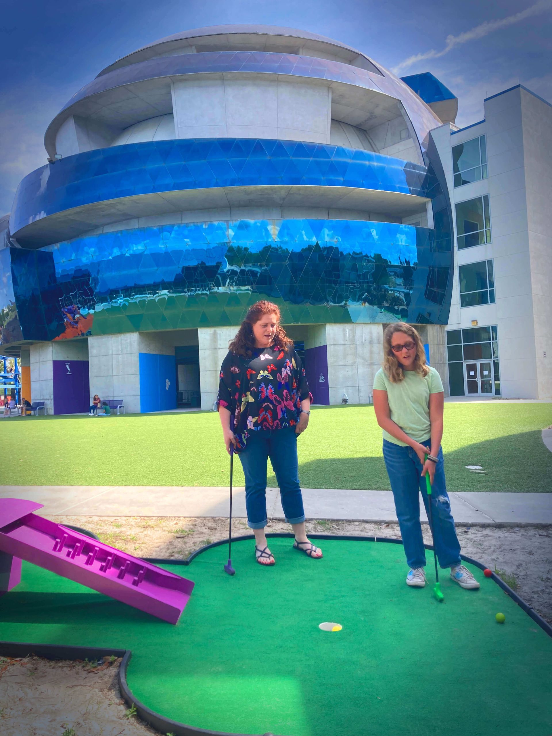 Two women are playing mini golf in front of a building
