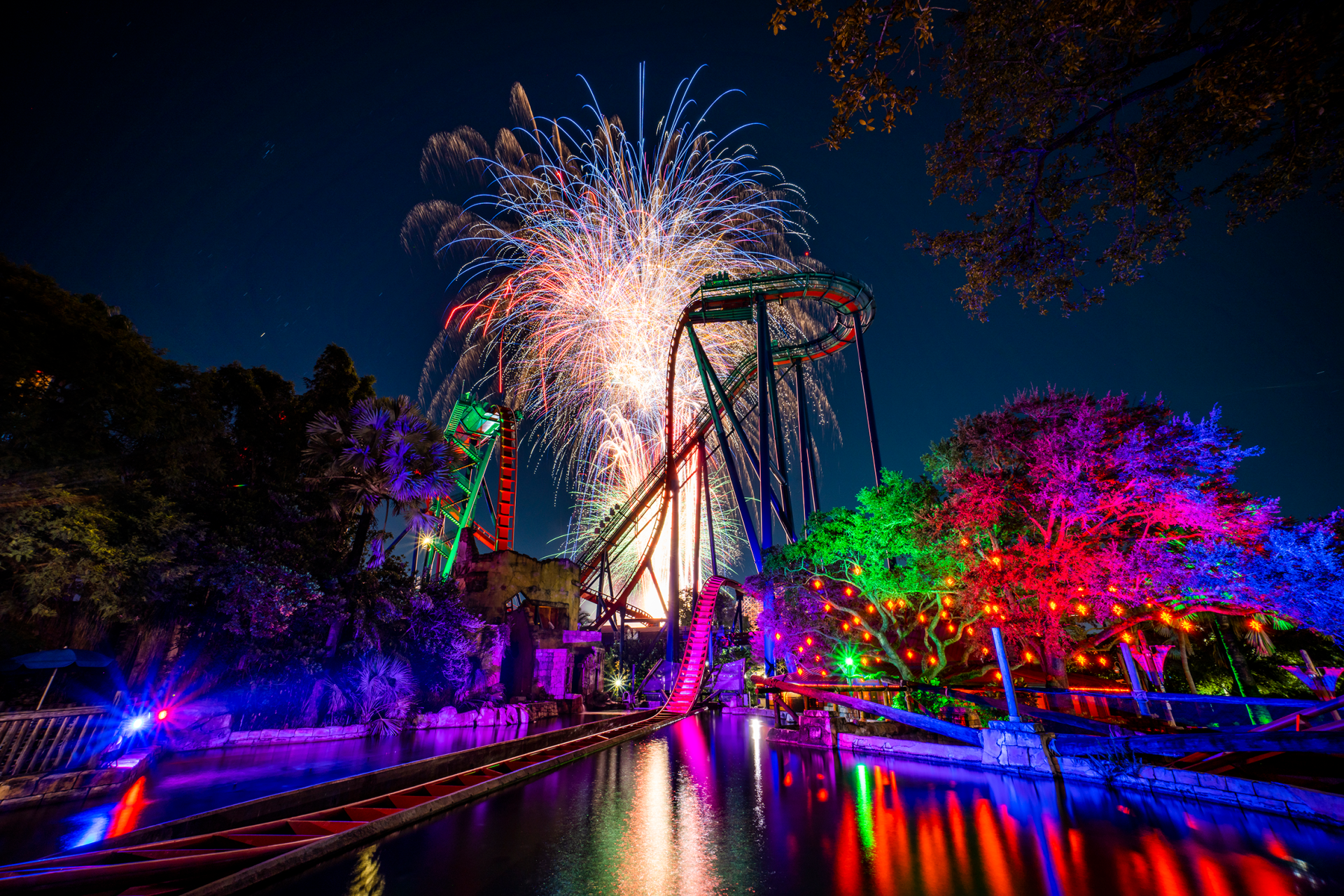 A roller coaster is lit up at night with fireworks in the background.