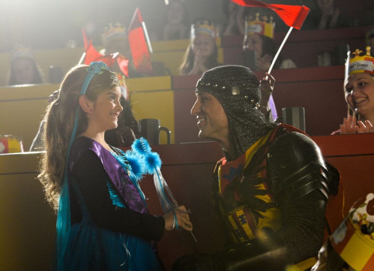 A man in a knight 's armor is shaking hands with a little girl in a blue dress.
