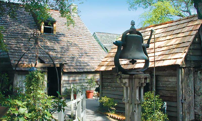 A large bell is sitting on a wooden post in front of a wooden house.