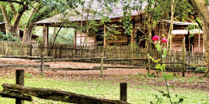 A wooden fence surrounds a log cabin in the woods.