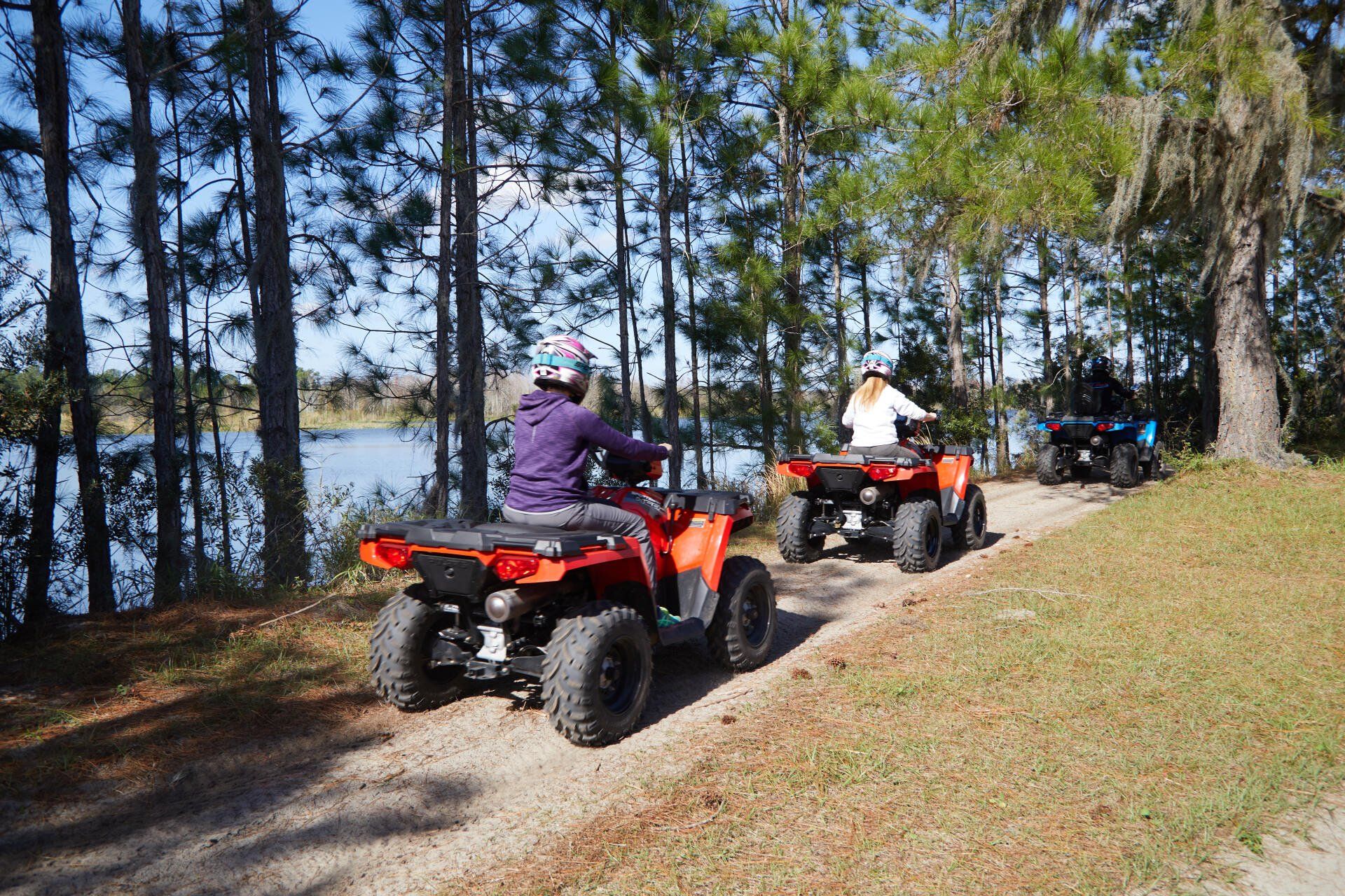Two people are riding atvs down a dirt road near a lake.