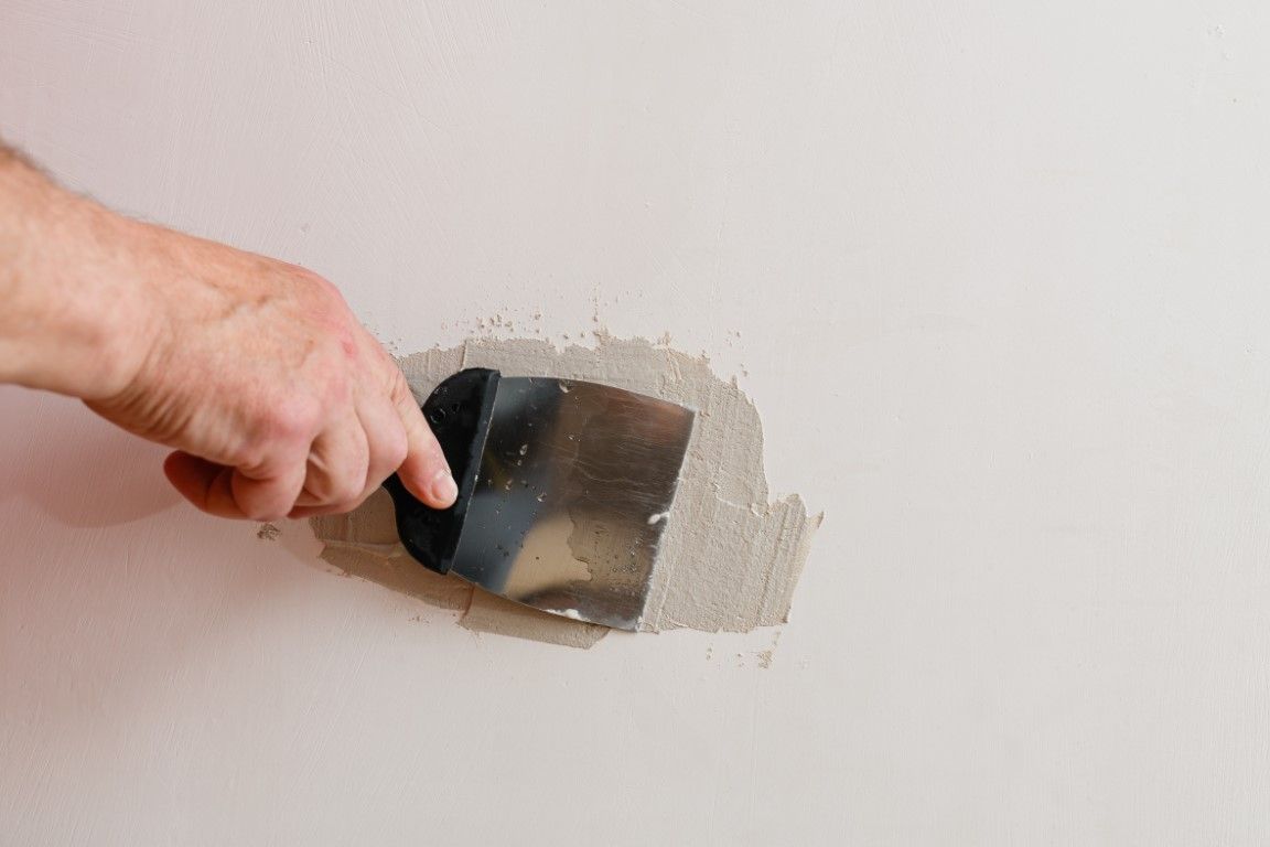 Hand using a putty knife to scrape plaster from a white wall hole