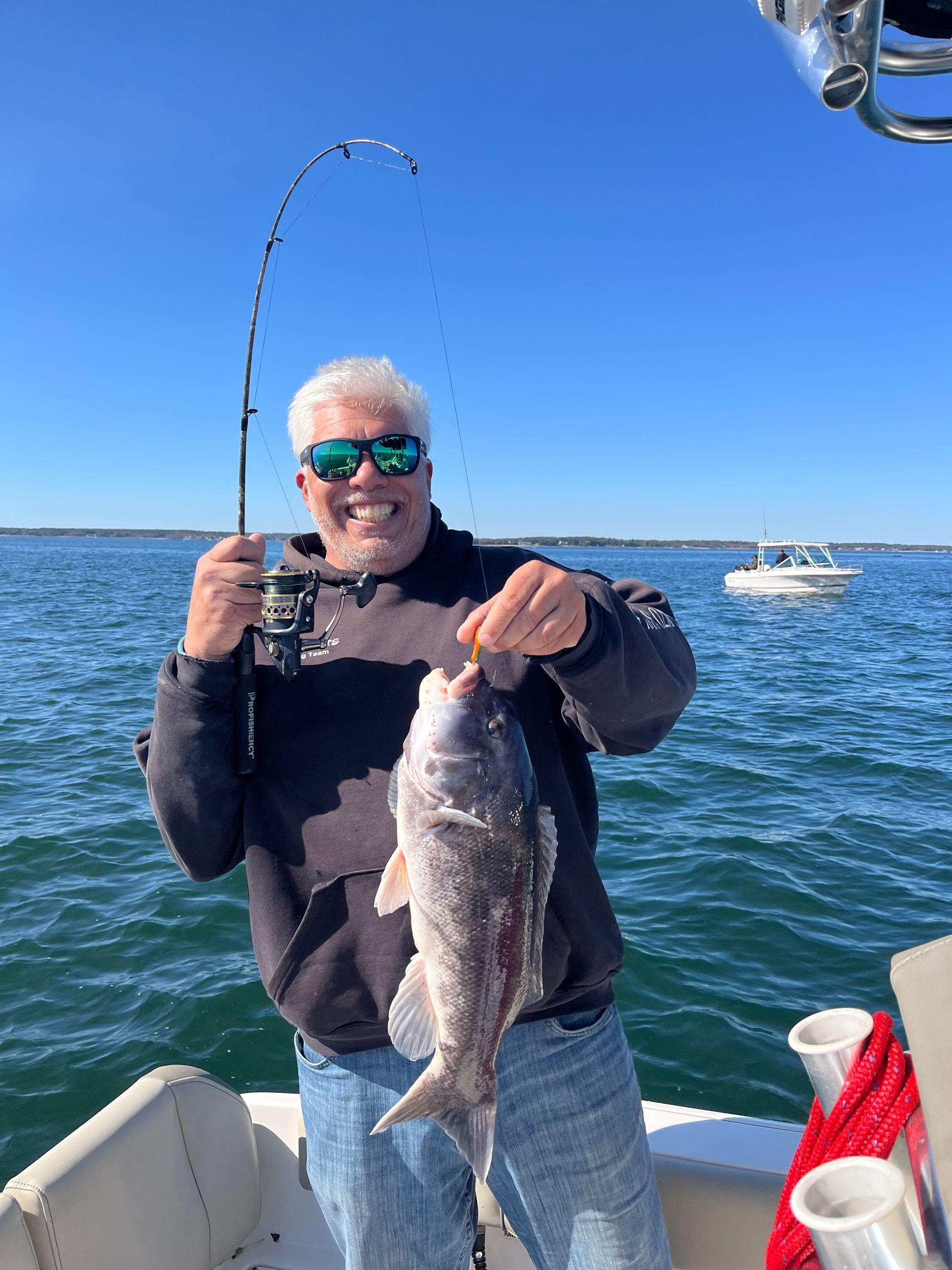 Man on boat smiling, holding up a fish he caught; blue sky and water in the background.