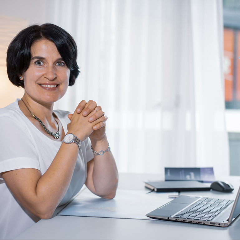 Frau lächelt an einem Schreibtisch mit Laptop, Notizblock und Telefon in einem hellen Büro.