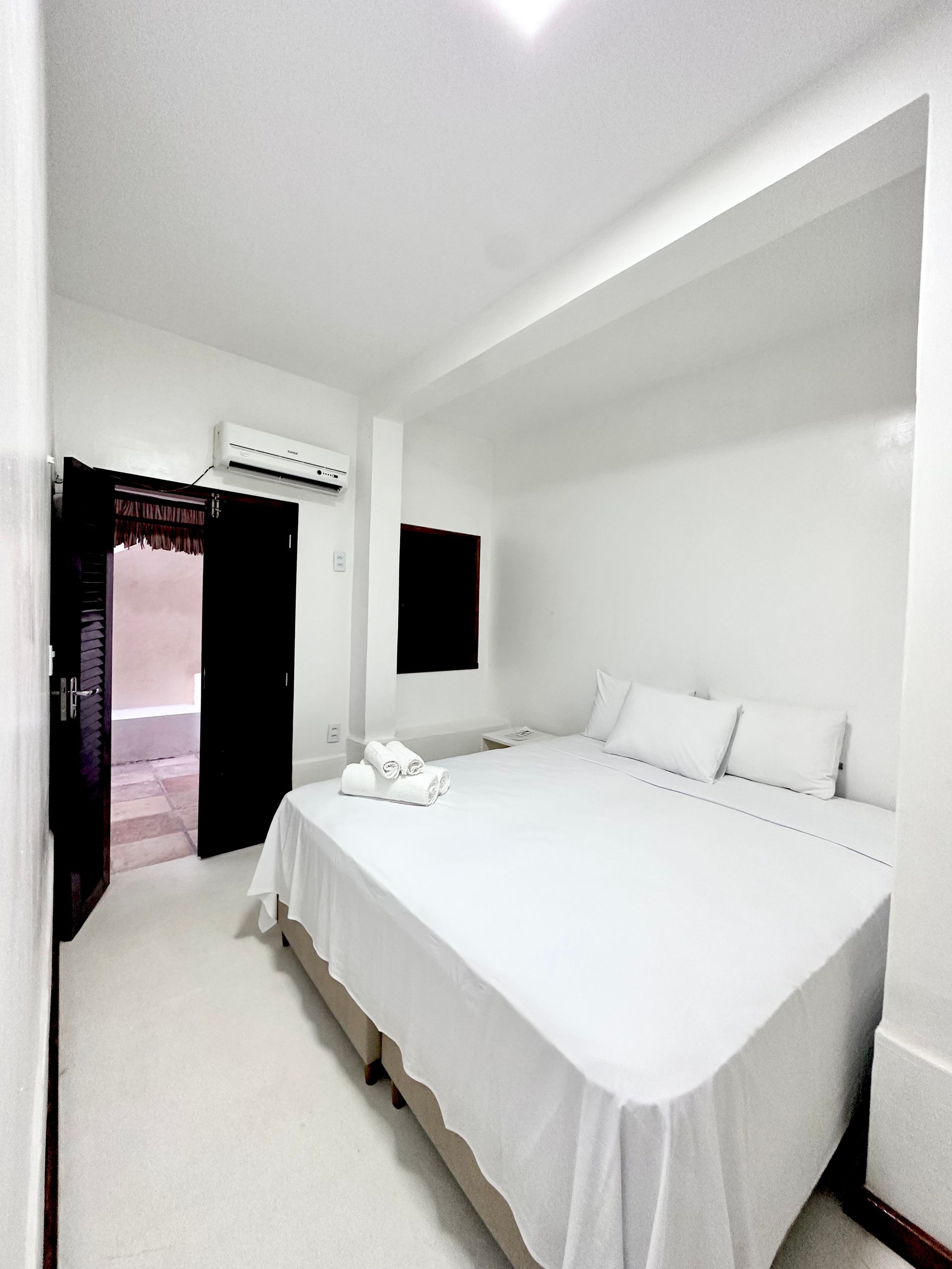 Quarto branco com uma cama grande, porta preta aberta e janela pequena.