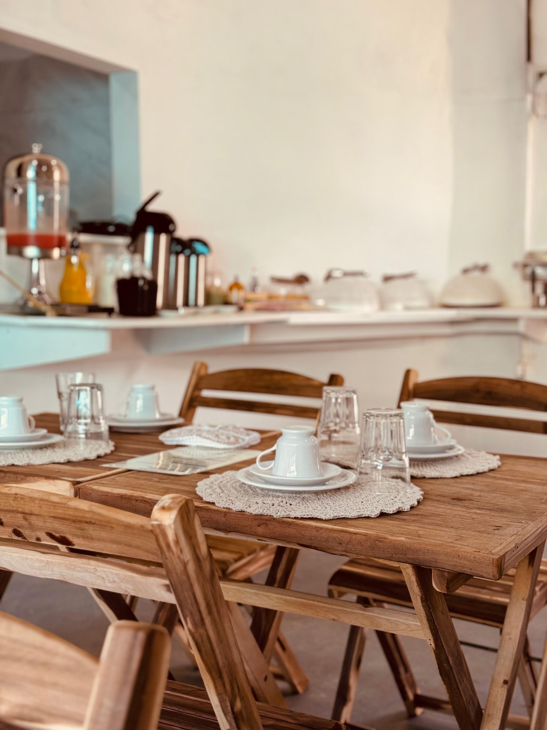 Mesa de madeira posta para o café da manhã com pratos brancos, toalhas de crochê e cadeiras em uma sala iluminada.