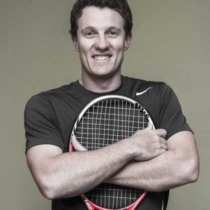Man smiling, holding a tennis racket with arms crossed; wearing a gray shirt, against a pale green background. Gary Innes.
