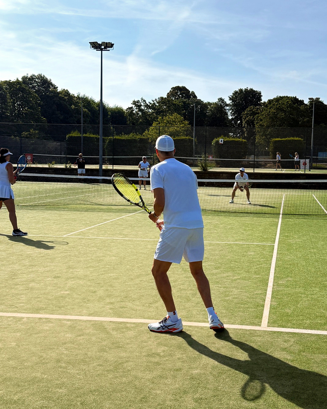 Tennis players on a sunlit outdoor court, with a player in white ready to return a shot.