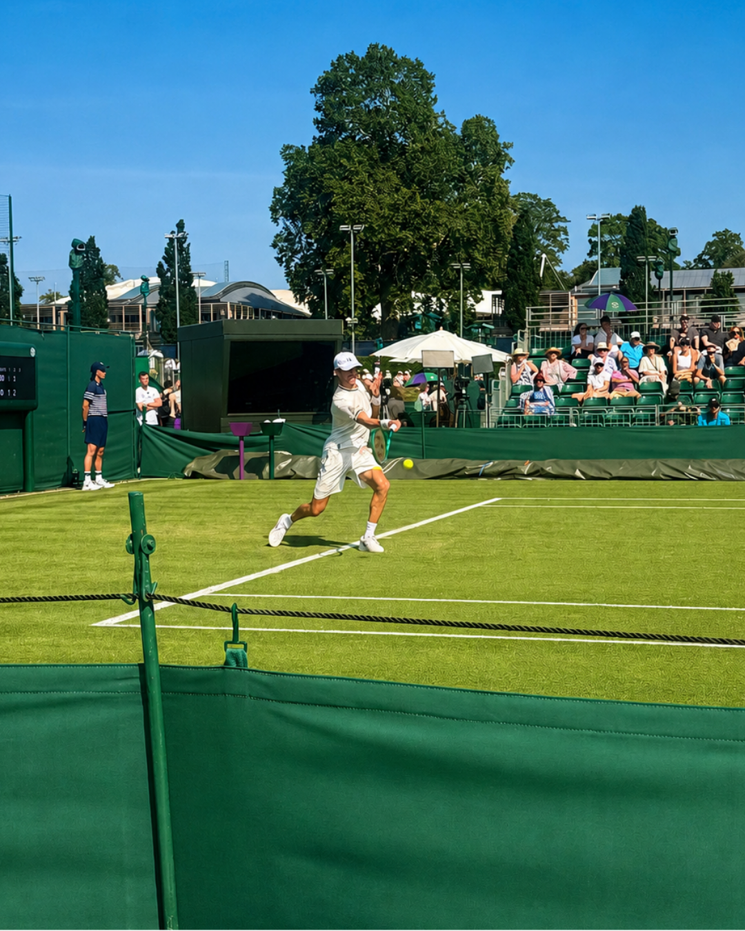 Tennis player in white and orange runs on a grass court during a match, with spectators behind the net.