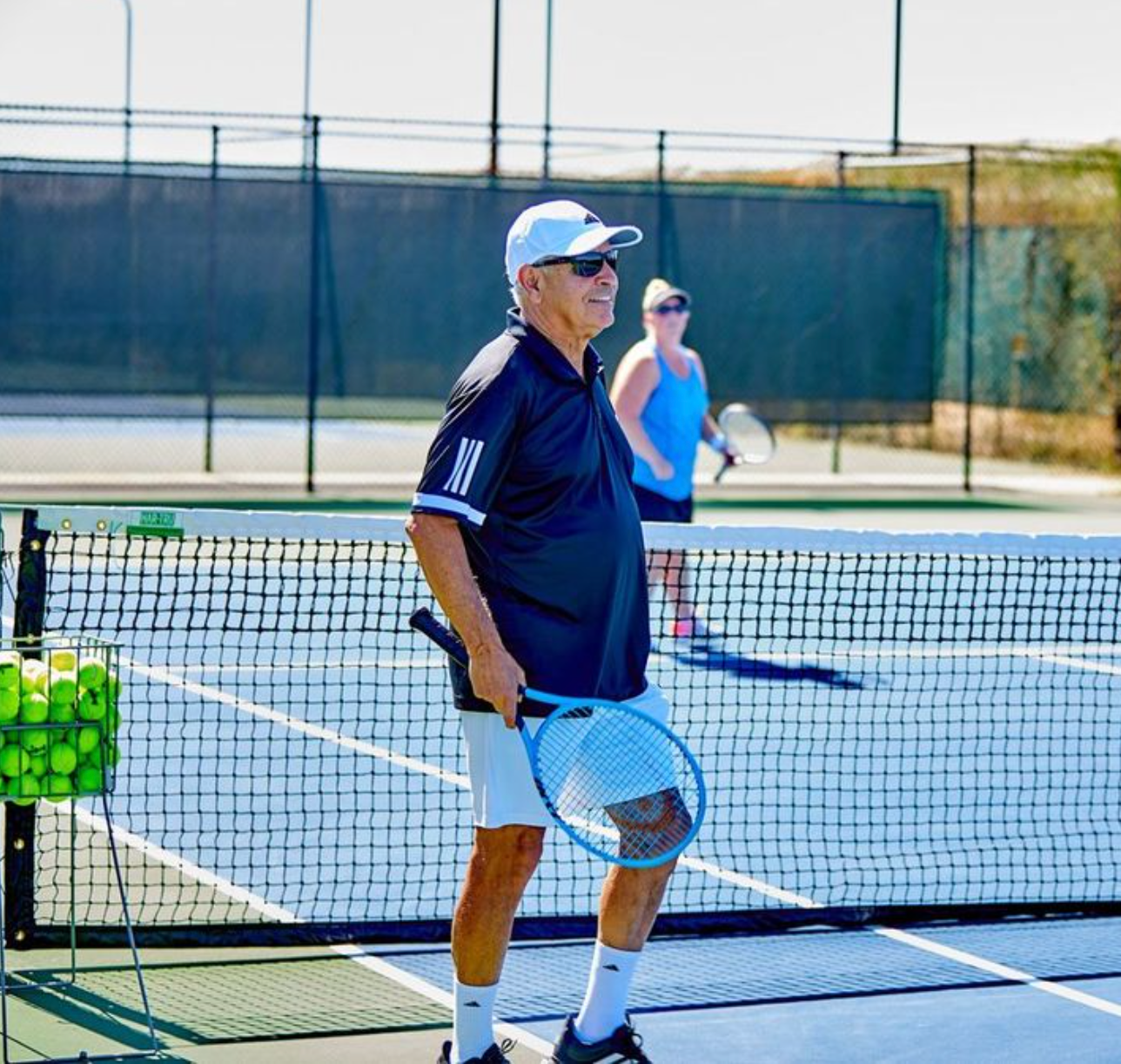 Man in sunglasses and white hat on tennis court, holding a racket. Another person in background.