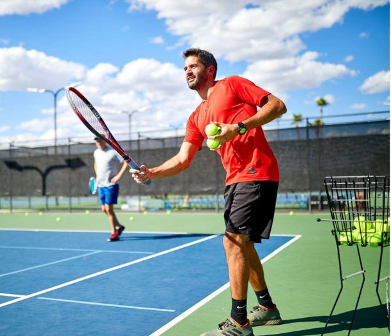Man on a tennis court holding balls, about to serve. He wears red shirt, black shorts. Another player in background.