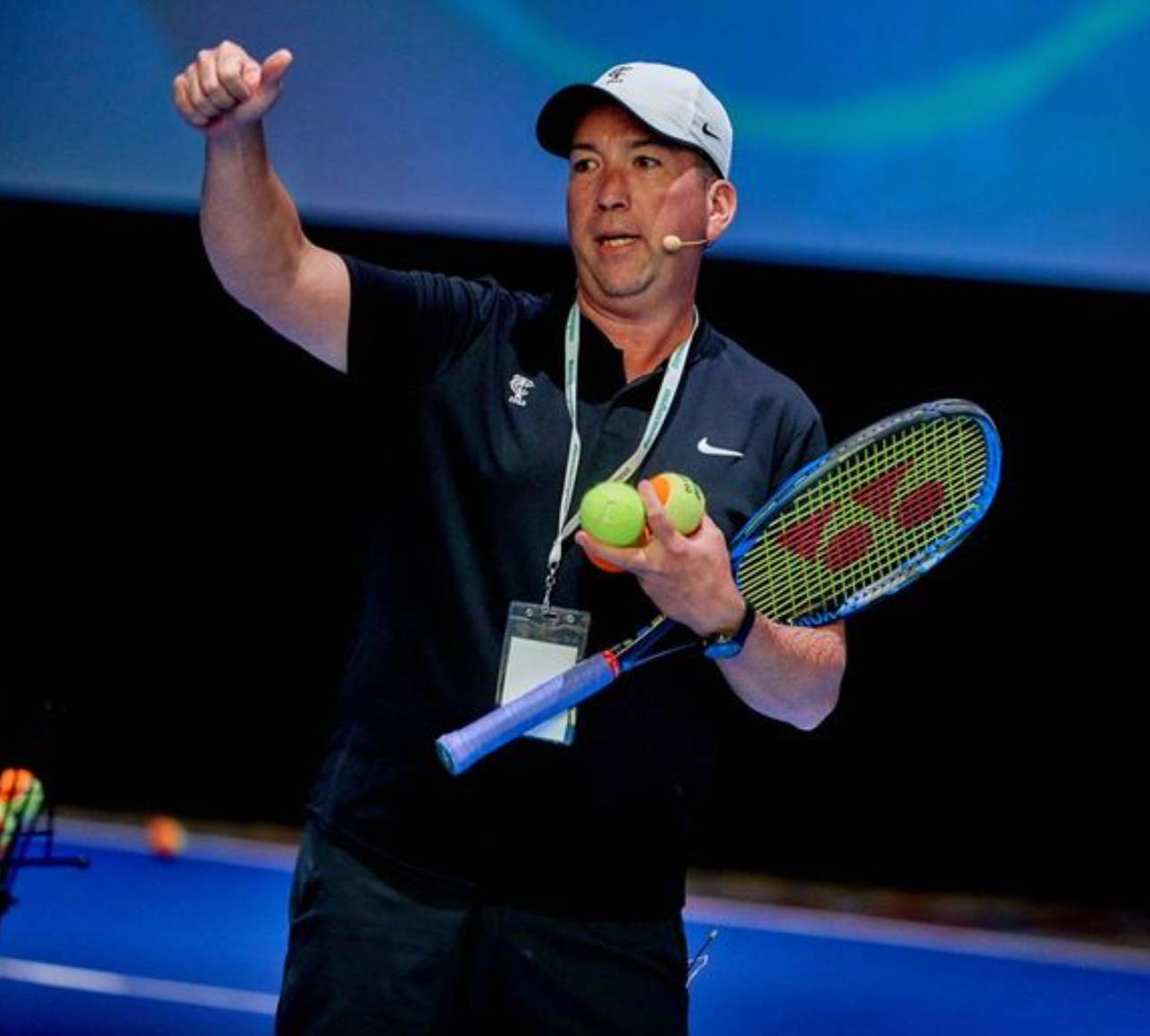 Man in tennis attire giving a presentation, holding balls and racket, gesturing with thumb up.