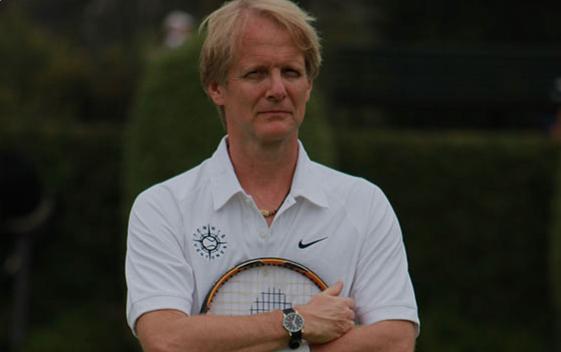 Man in white shirt holding tennis racket on a grassy area, looking off to the side.
