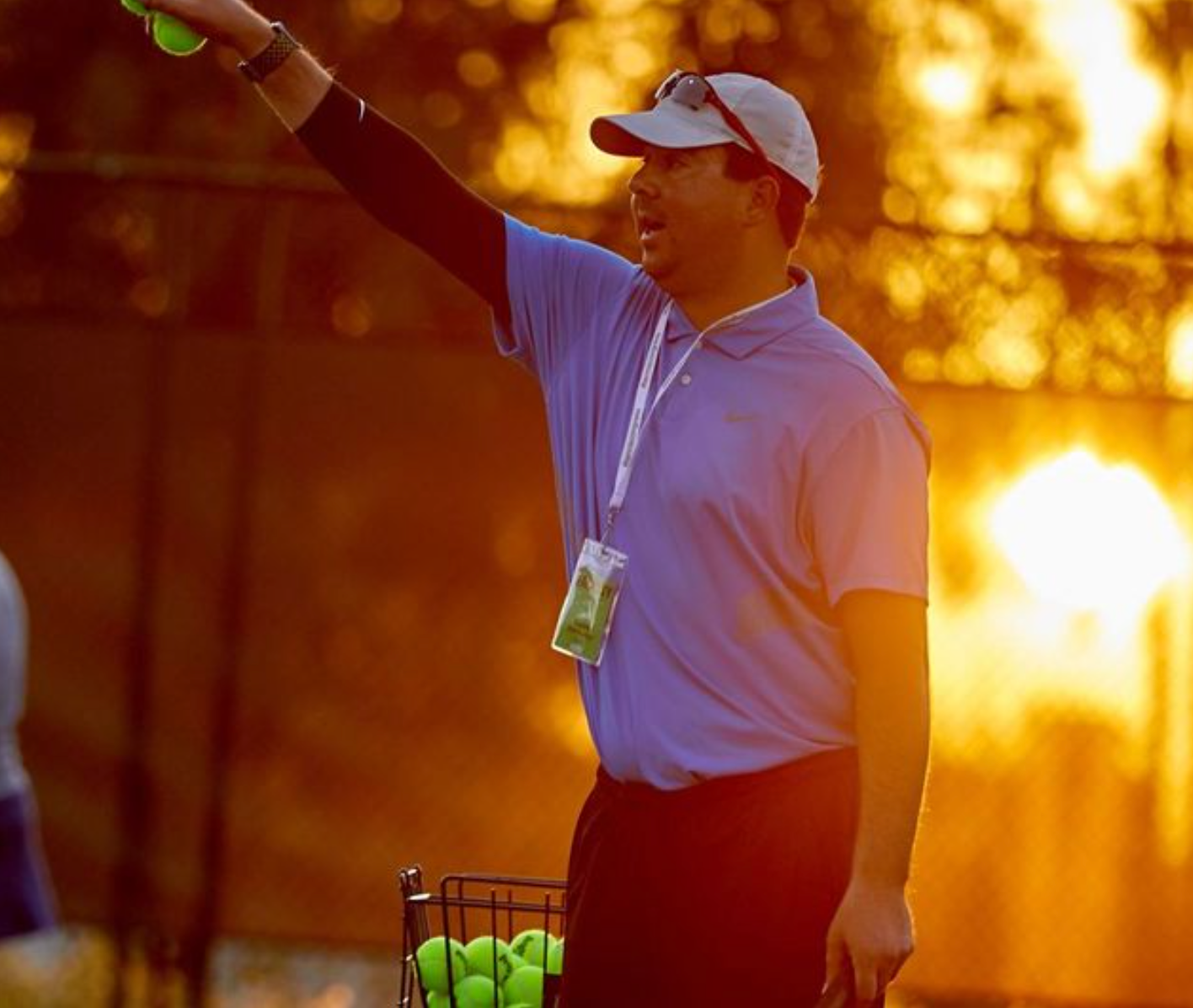 Tennis coach in a blue shirt with a cap, holding a ball, outside on a sunny day.