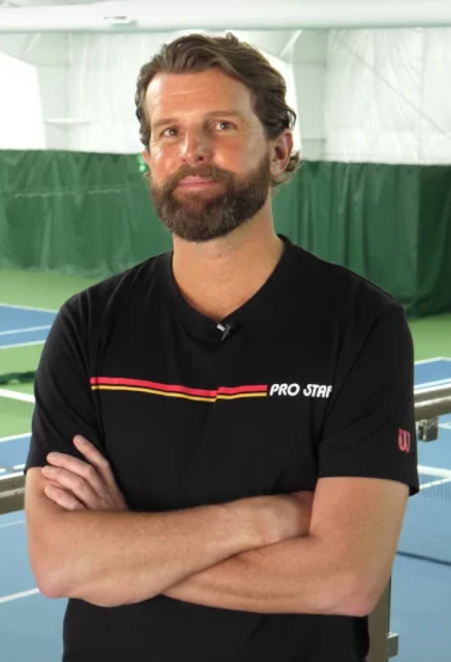 Man with beard in black shirt, arms crossed, on a tennis court.