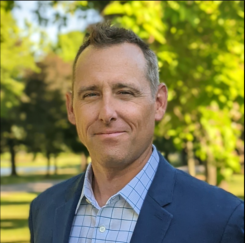 Man in a blue suit smiling, outdoors with trees in the background.