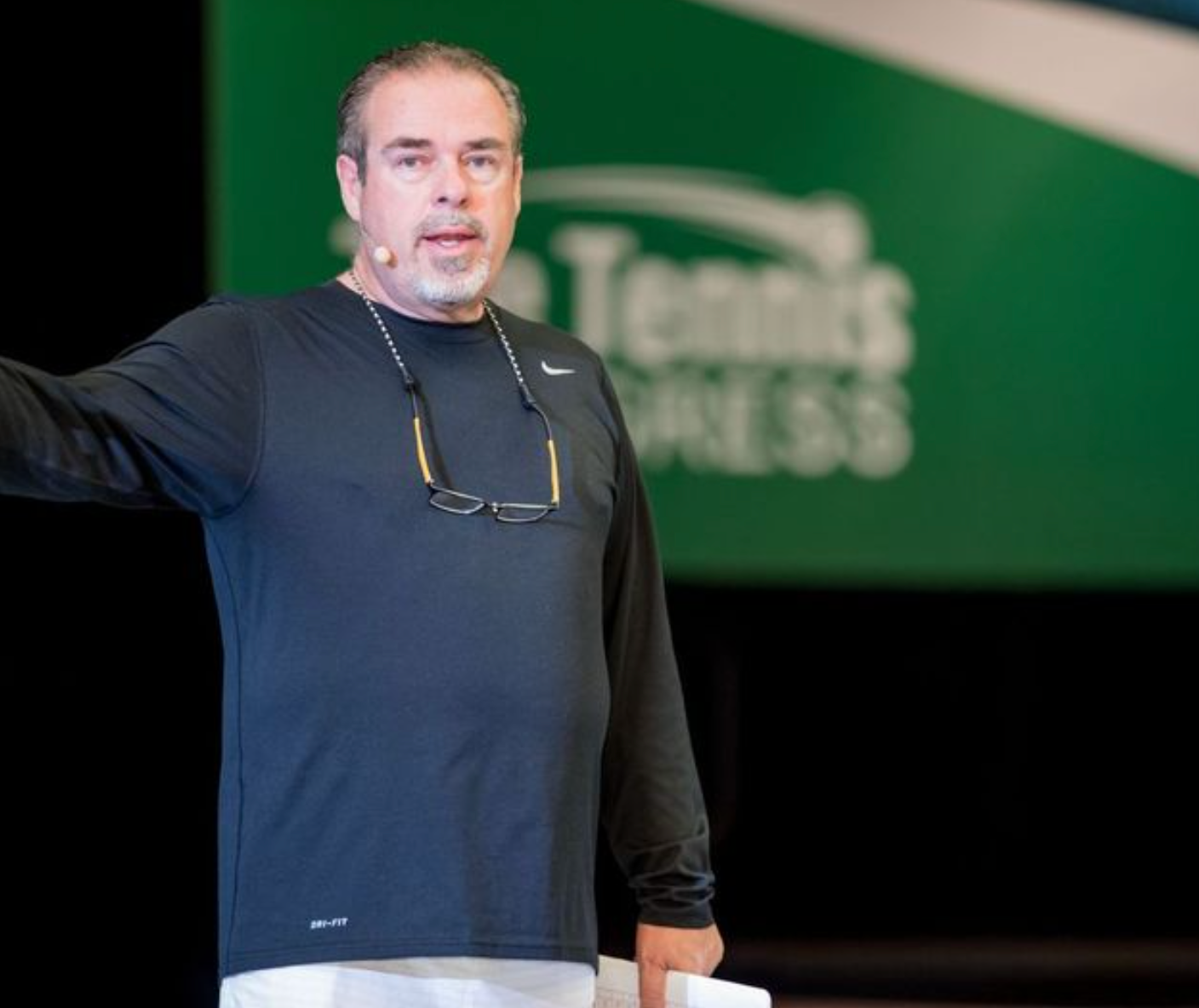 Man in black shirt with glasses, speaking on stage at a Tennis Express event.