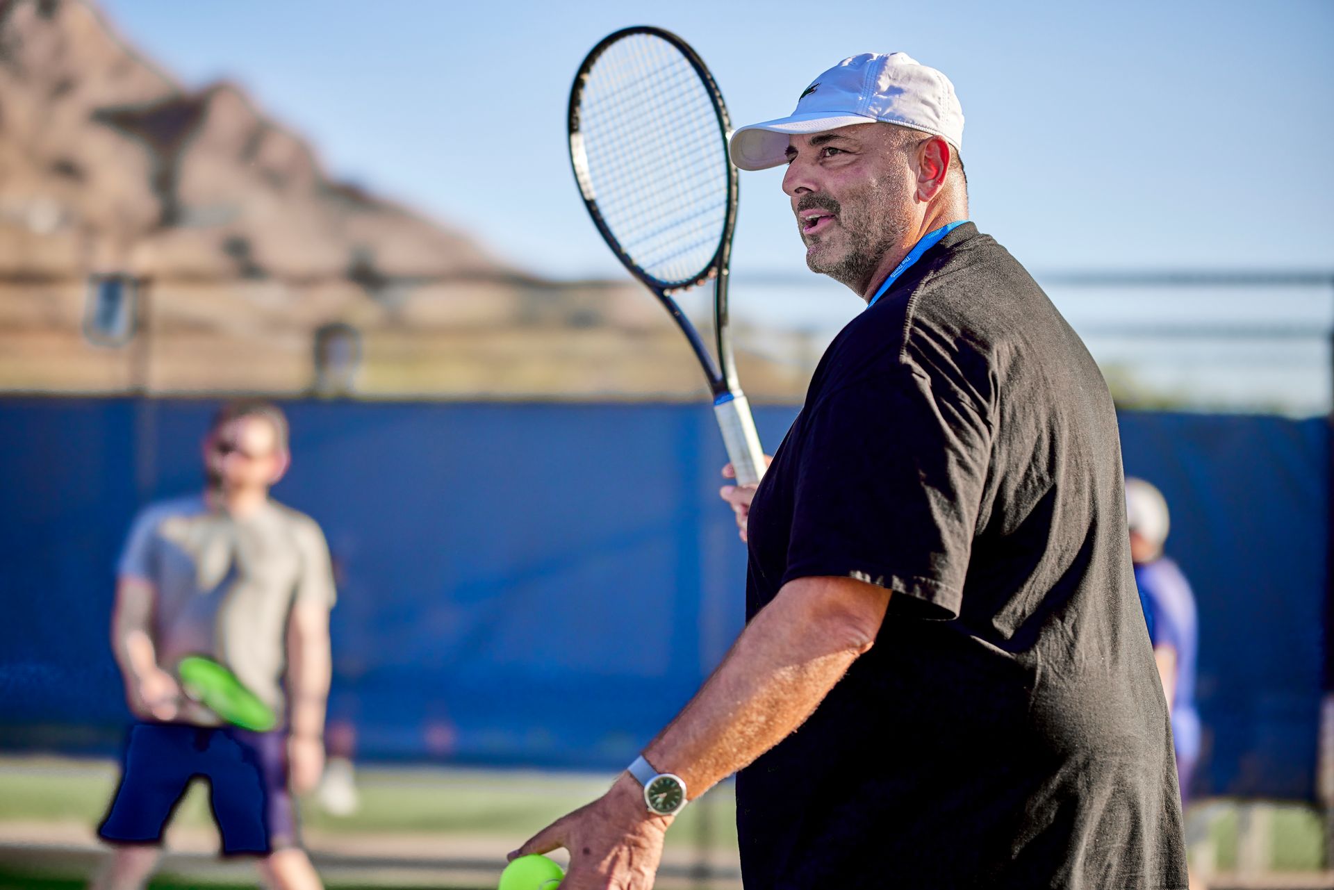 Man on a tennis court holding a racquet, looking to the side. Wearing a white hat, black shirt.