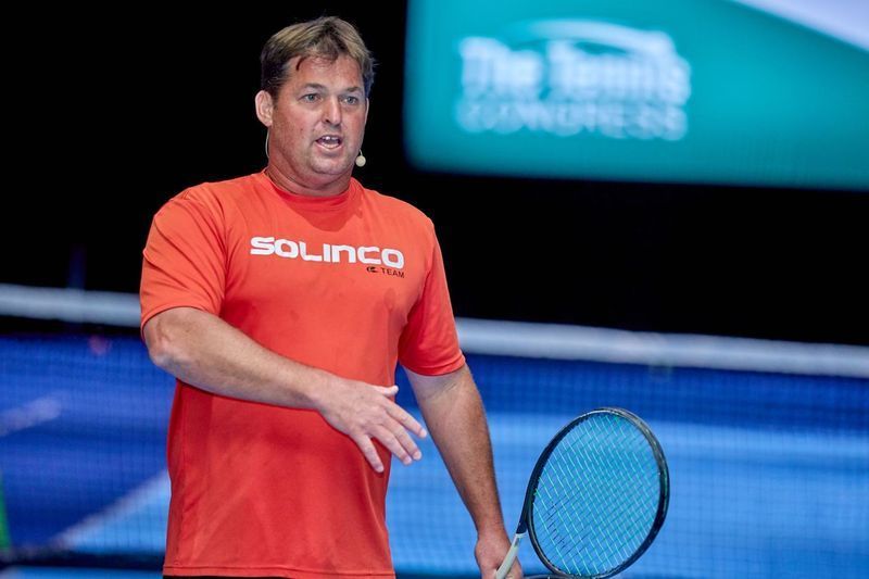 Man in red shirt holding tennis racket, speaking at 