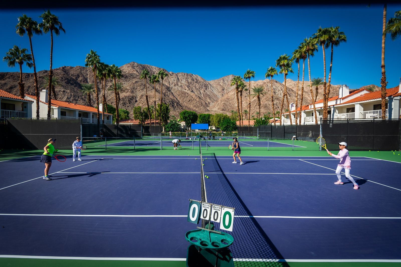 Tennis doubles match on a purple court with players, scoreboard, palm trees, and mountains in the background