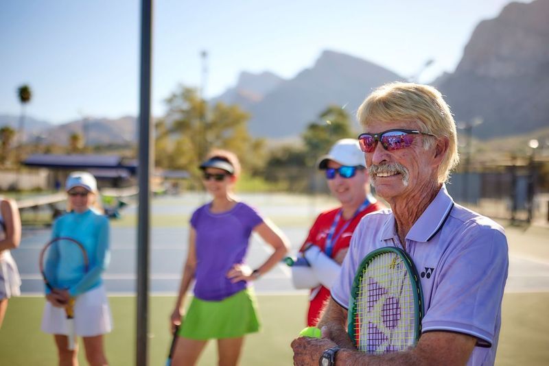 Man with sunglasses and tennis racket talking to a group on a court with mountains in the background.