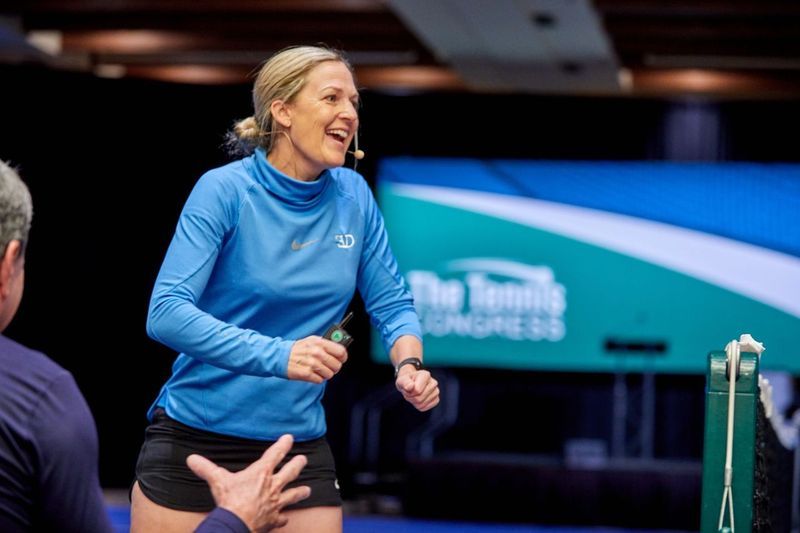 Woman in blue shirt laughs, gestures near tennis net; 